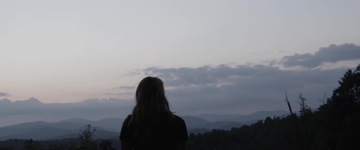 A blonde woman walking through a field with mountains and a cloudy sky in the background