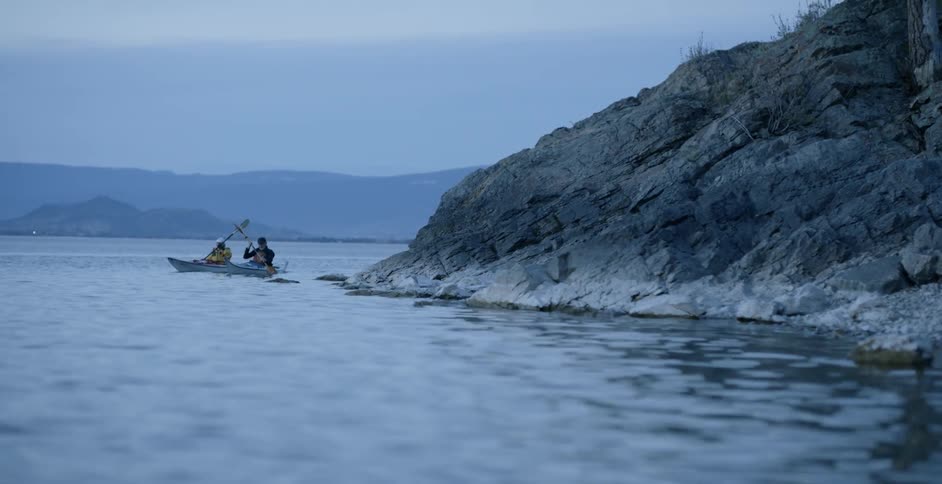 Two people kayaking at dusk next to a hill