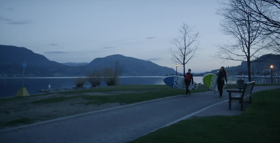 Two women are carrying their paddle boards along a pathway at dusk