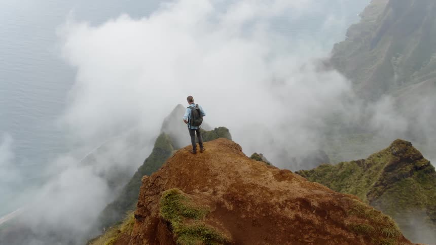 A hiker is standing on a ledge above a mountainous coastline in daylight