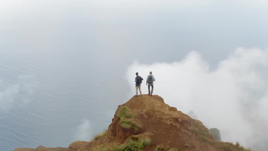 Two men stand on a mountain and look around at the clouds