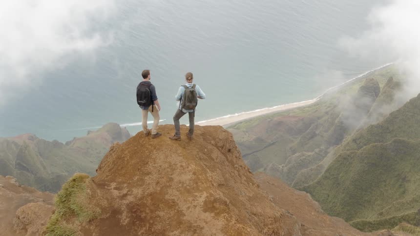 Two men are standing on a ledge overlooking coastal mountains in daylight