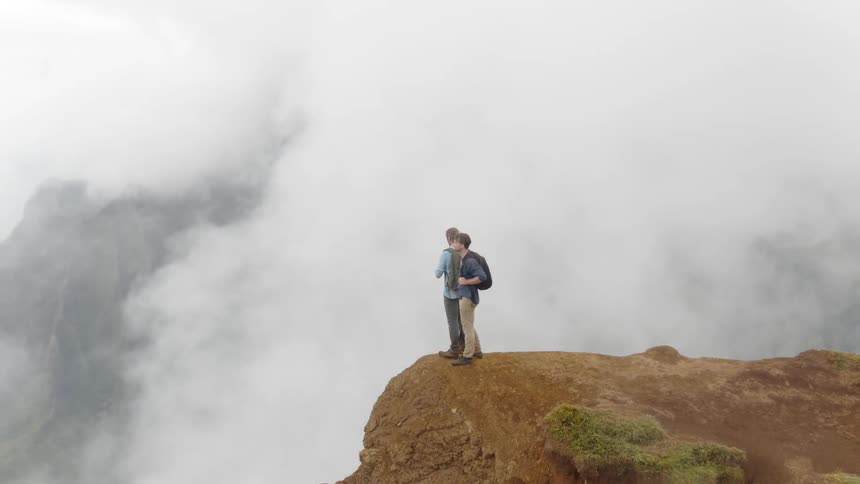 Two men stand on top of a mountain and look out at the clouds