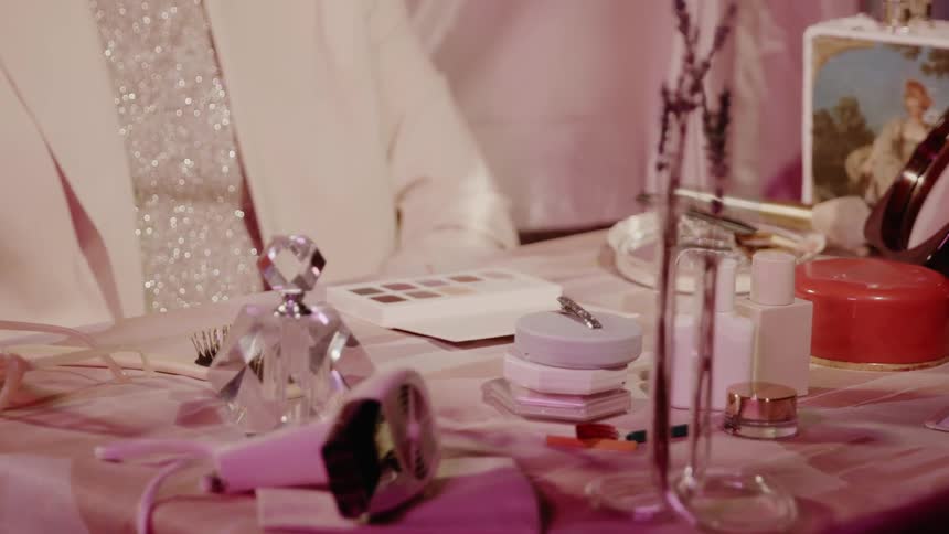 A woman sits at a dressing table with cosmetics