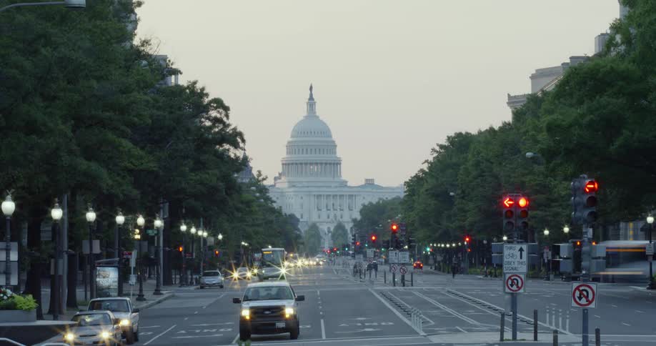 Cars drive on a street in front of a capitol building at sunrise