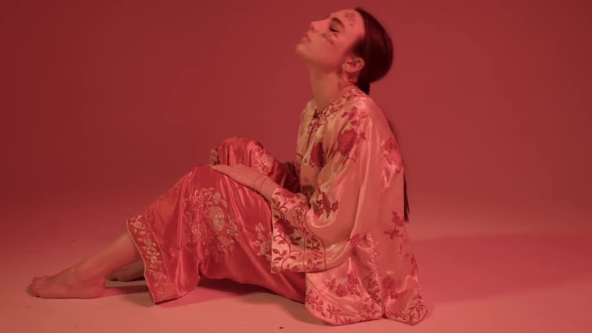 Woman wearing robes and face paint and sits and rocks in red illuminated light