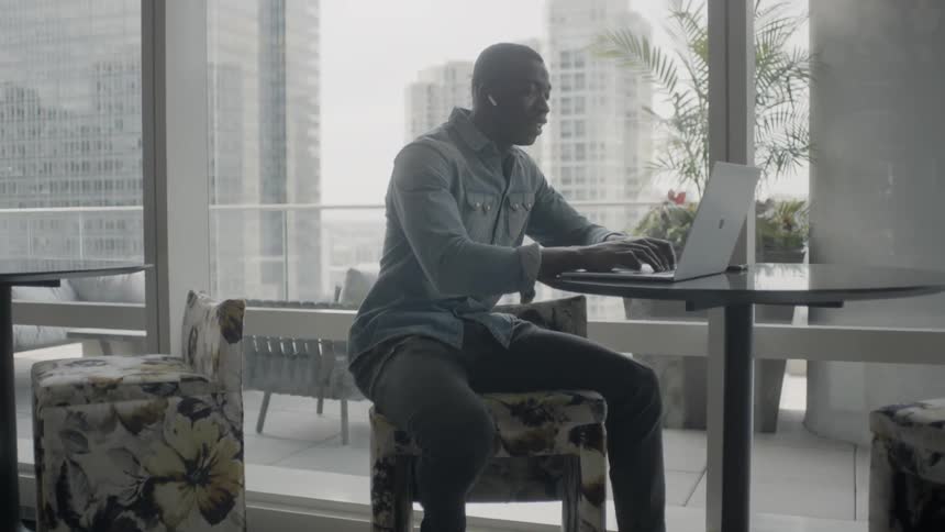 A man is sitting at a table in a high-rise building working on a laptop in daylight