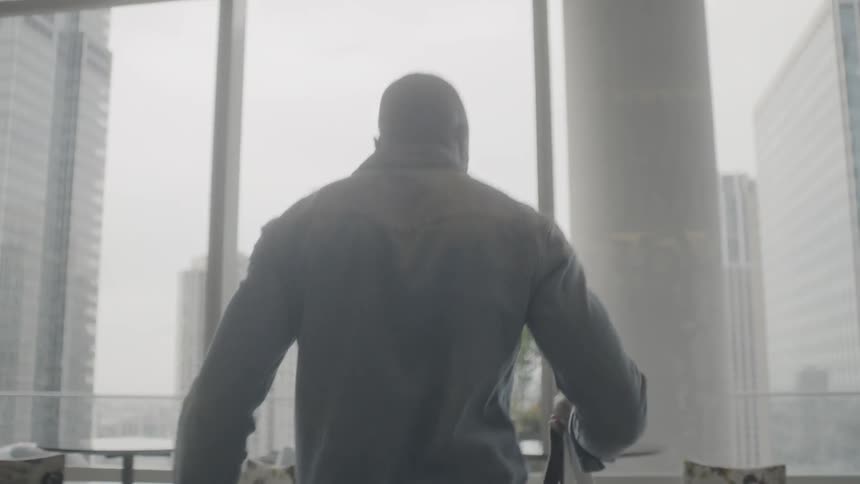 A man is walking towards a table, sitting down, and pulling out a laptop inside in daylight
