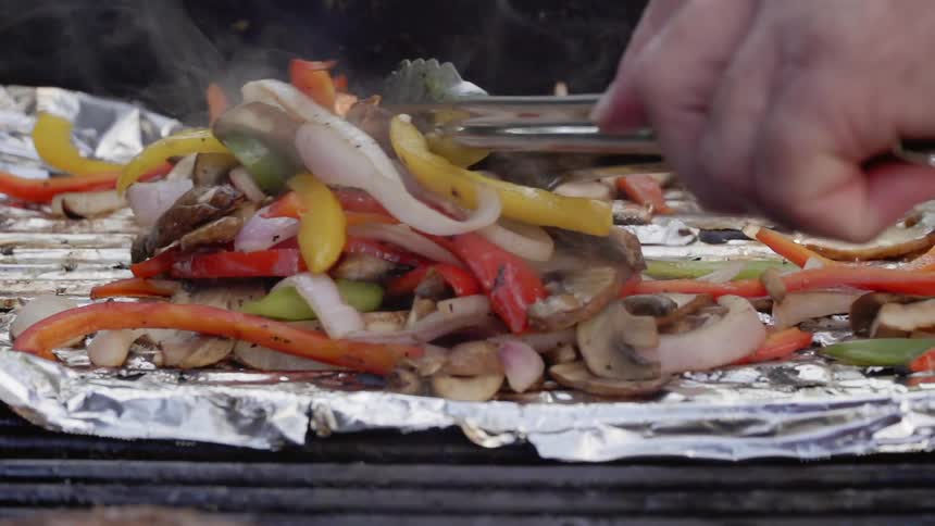 A person uses tongs to spread out vegetables cooking on foil on a grill