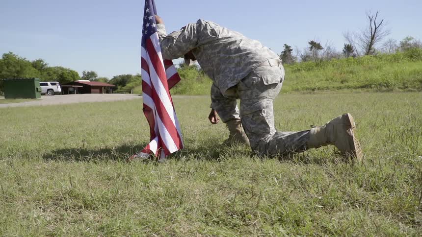 A soldier is leaning on a flag as he rises from kneeling on the ground outside in daylight