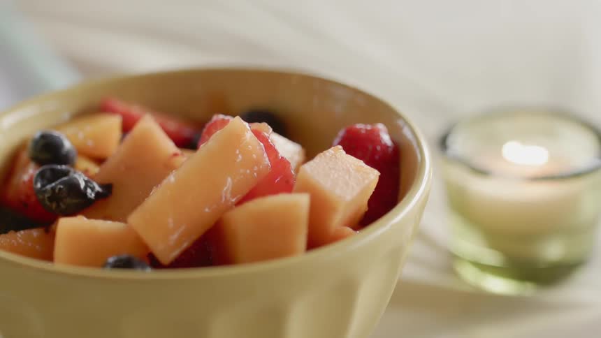 A candle is burning near a bowl of fruit inside in daylight