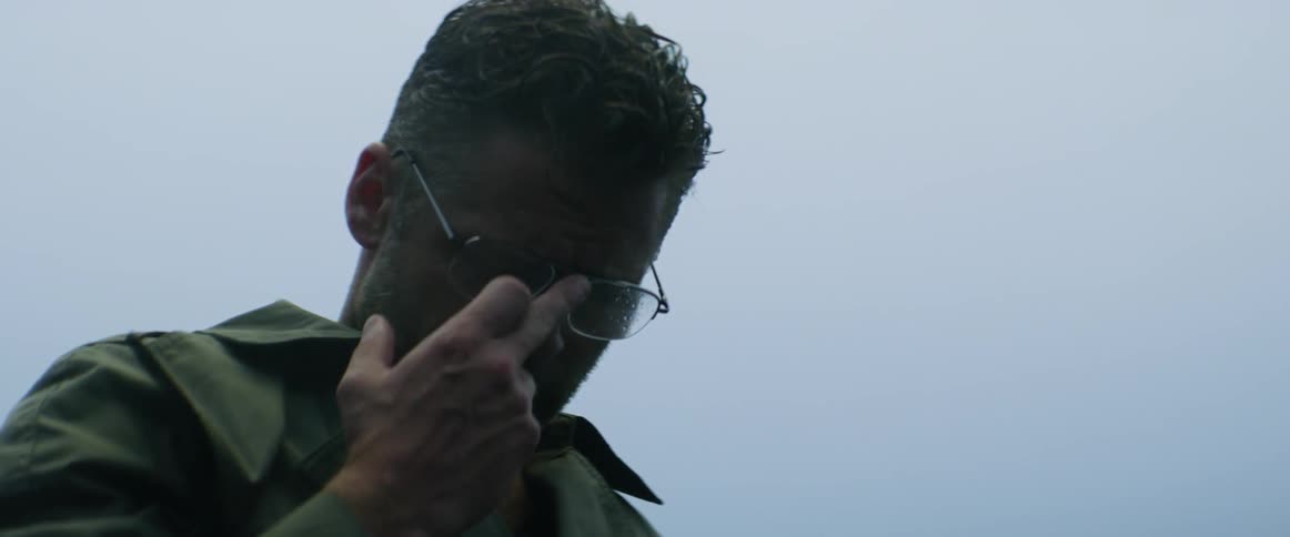 A man is shrugging and pushing up his glasses as he stands outside on a rainy day