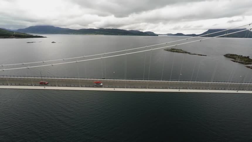 Vehicles travelling over a bridge suspended over water with mountains and low-lying clouds in the background