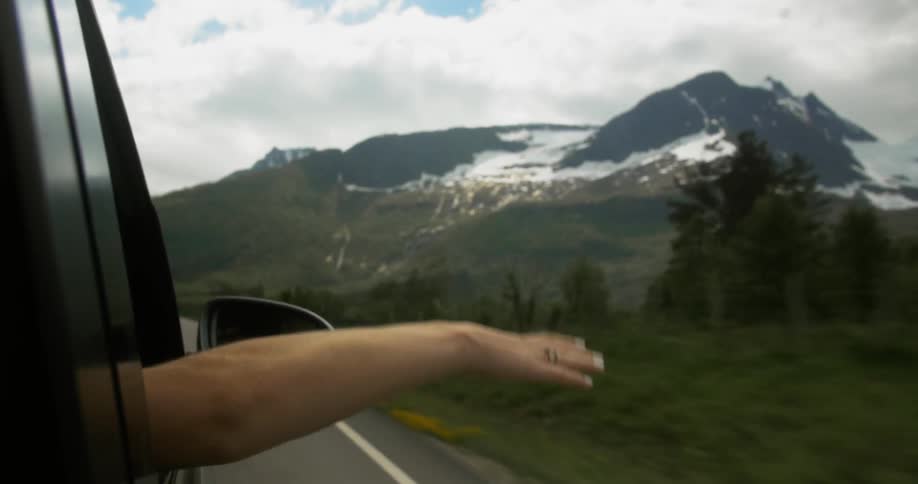 A person with their arm hanging out of the window as they travel along a road towards snow-capped mountains