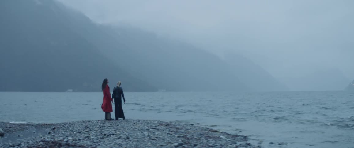 Two women with an umbrella running and playing near a lake and mountains