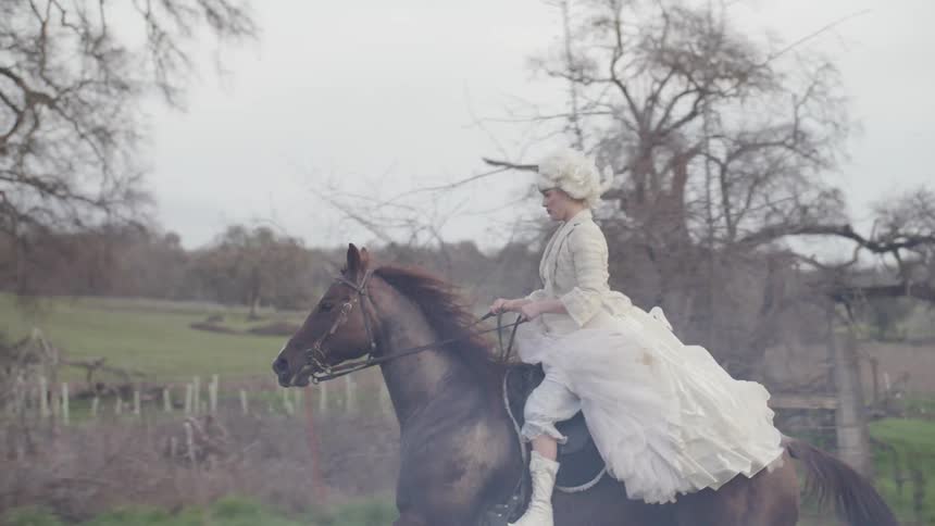 A woman wearing a period costume is riding a horse in a country setting in daylight
