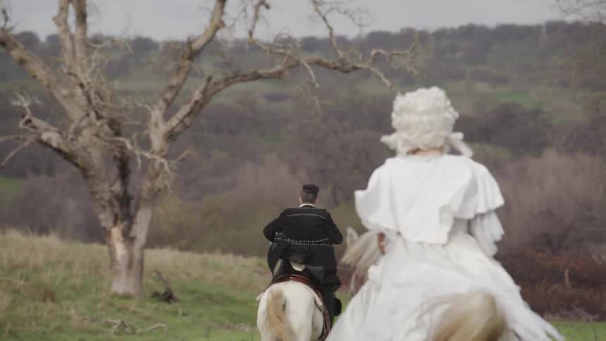 A man and two women in old fashioned clothes riding horses during the day