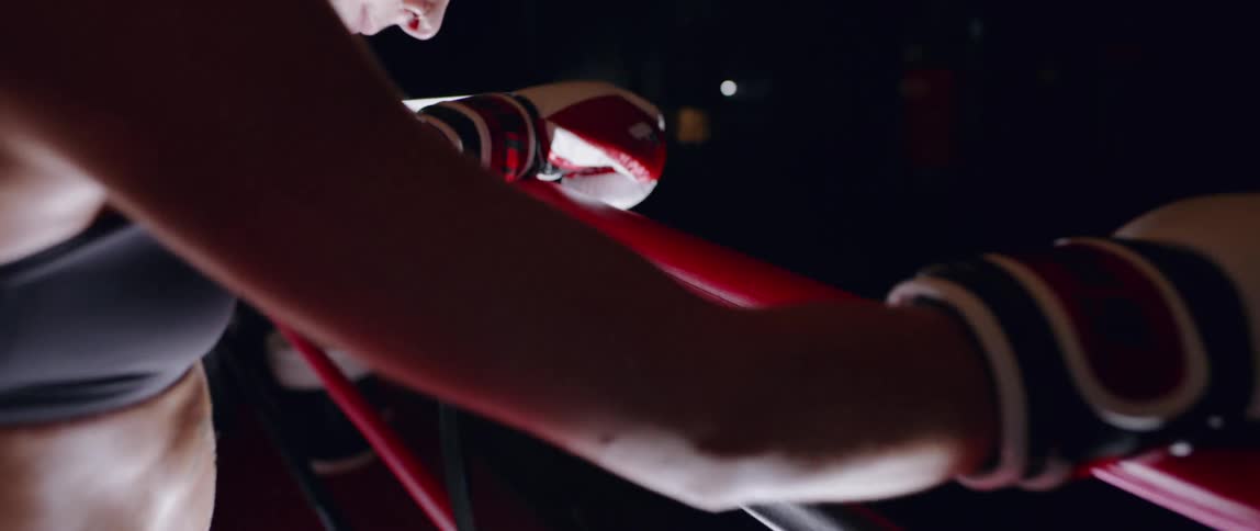 Woman wearing boxing gloves leans over ropes in a boxing ring