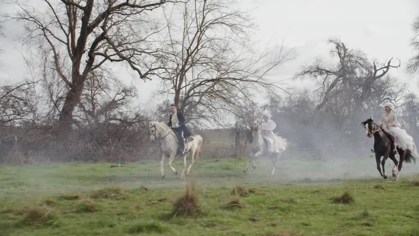 Several people ride horses across a field on a foggy day