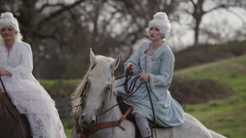 Women in white wigs and gowns sit on horses in a field