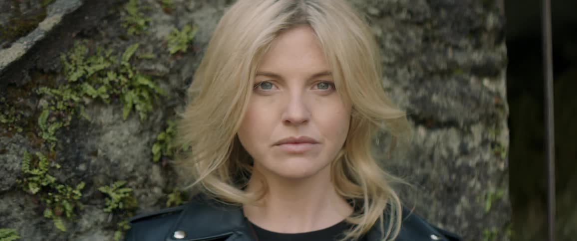 A woman with blond hair leans against a rock wall dotted with ivy