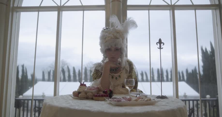 A woman in a tall white wig sits at a table with plates of food and wine