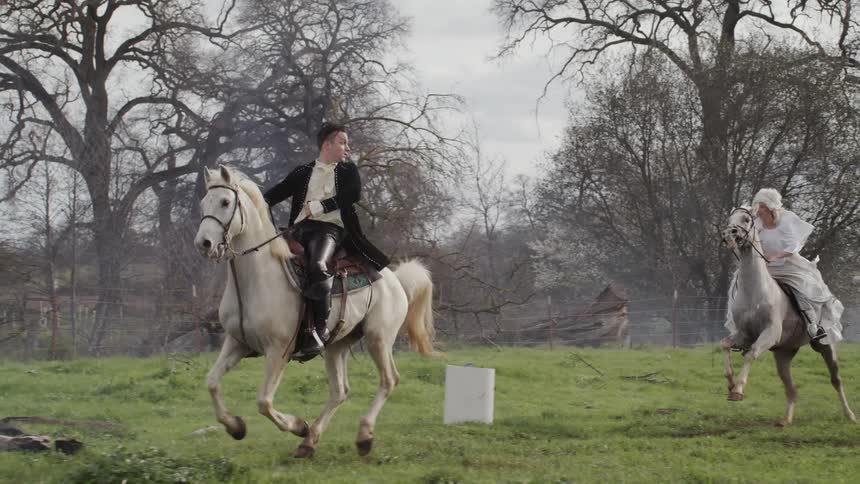 Man and woman ride white horses in a field on a cloudy day