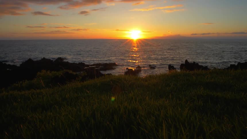 A sunset over the ocean seen from a grassy and rocky shore