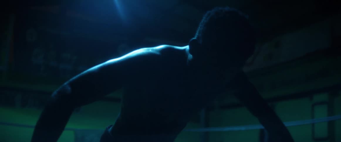 Shirtless man stands in a boxing ring leaning over the ropes