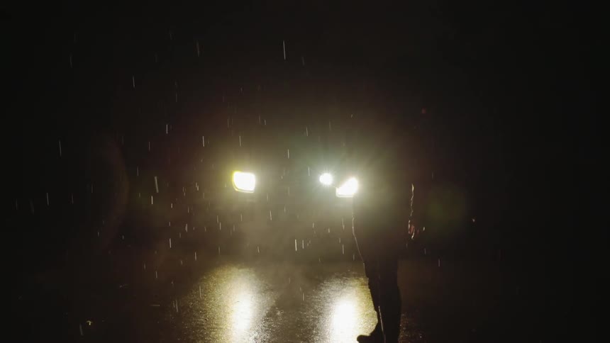 Man holds a flashlight as he stands in the rain in front of a car