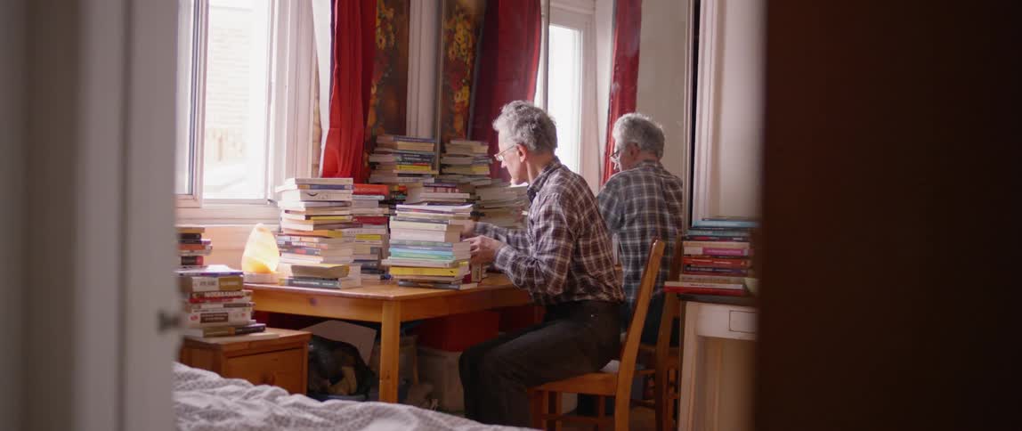 A man sits at a desk covered with stacks of books