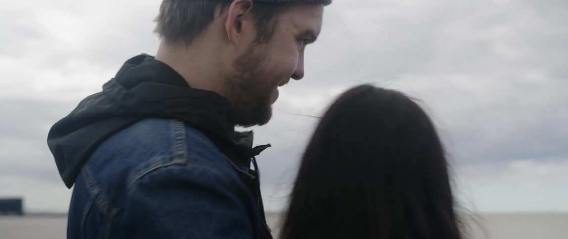 A man is kissing his partner as they stand outside under cloudy skies