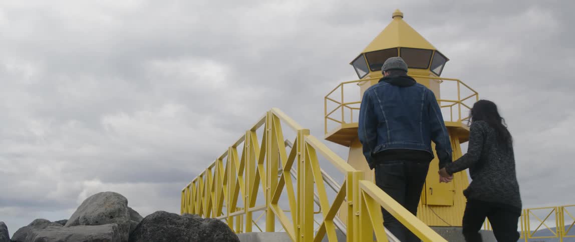 A couple holds hands as they walk up the steps near a yellow lighthouse