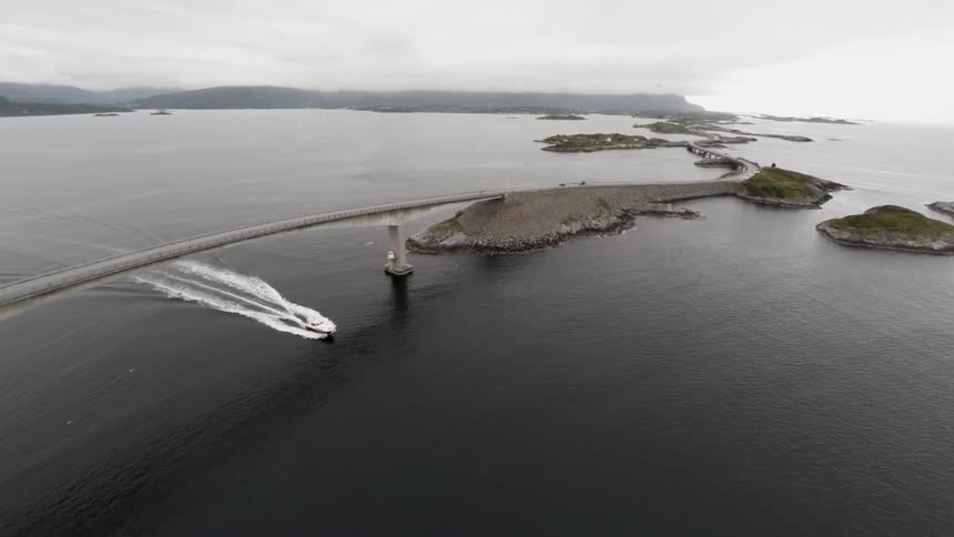 A bird's-eye-view of a boat traveling on a body of water that has small islands dotting it and cars travelling on a bridge that spans the water