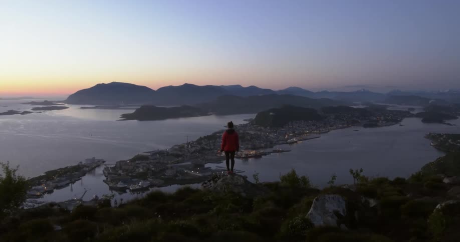 A woman stands on a rock overlooking a coastal town at dusk