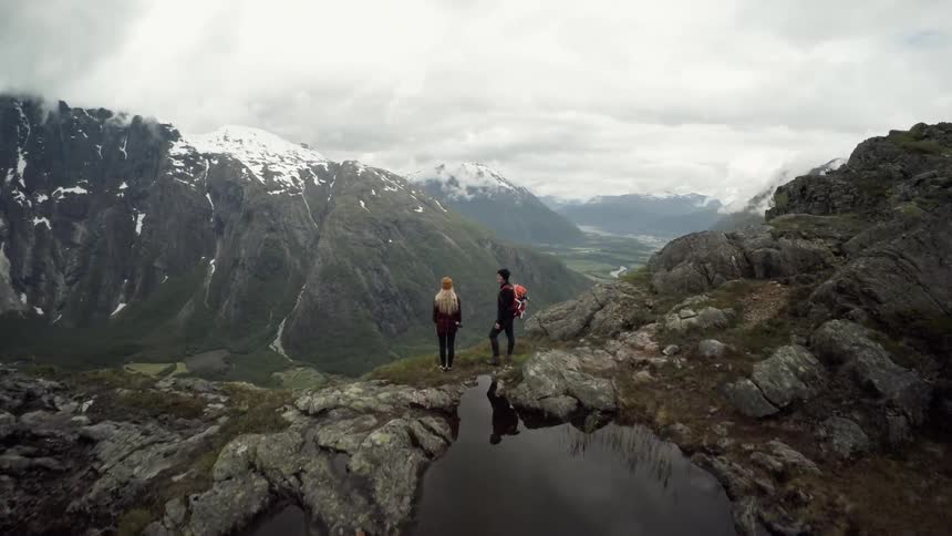 Two people stand at the edge of a small pond near the edge of a mountain and look at the valley below