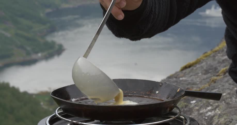 A man cooking pancakes on a camping stove with a body of water and cliffs in the background