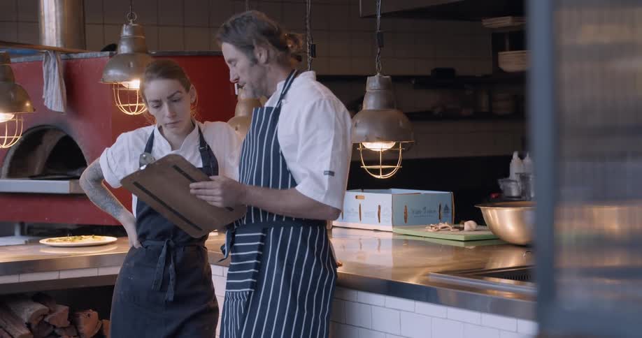 Man and woman stand in a restaurant kitchen talking and looking at a clipboard