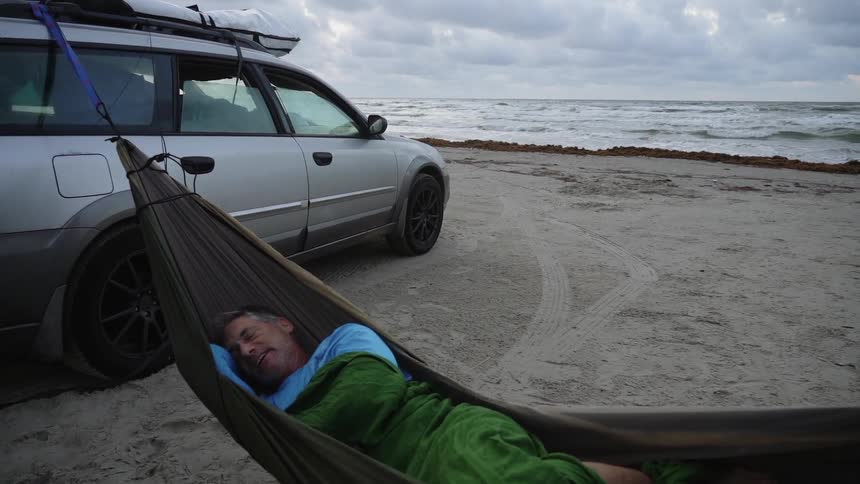 Man sleeps in a hammock connected to his vehicle on the beach