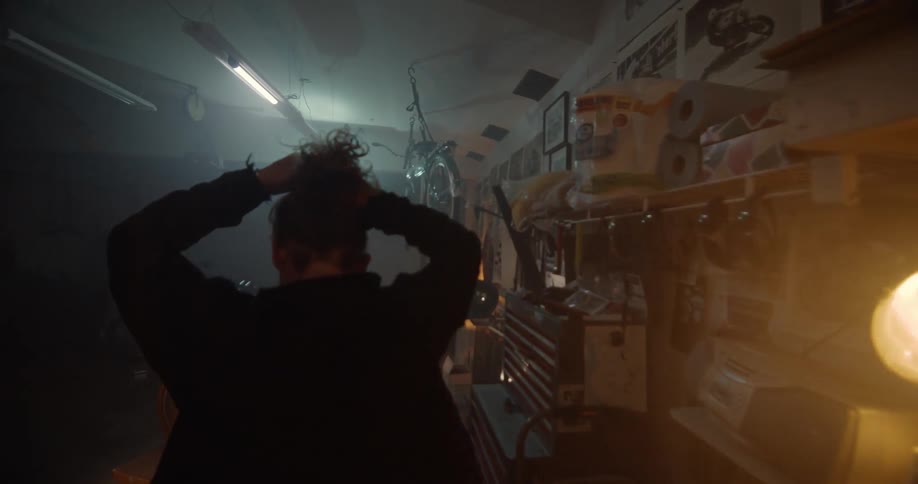 A man is pulling back his hair as he walks through a garage filled with motorcycles and gear