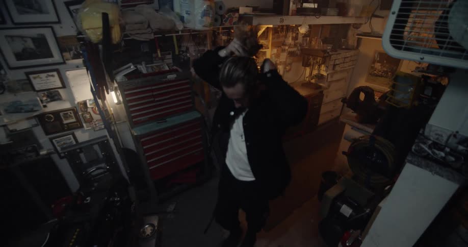 A man is pulling back his hair as he walks through a garage toward a motorcycle