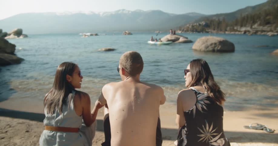 Three friends are sitting and talking on a lakeshore on a sunny day