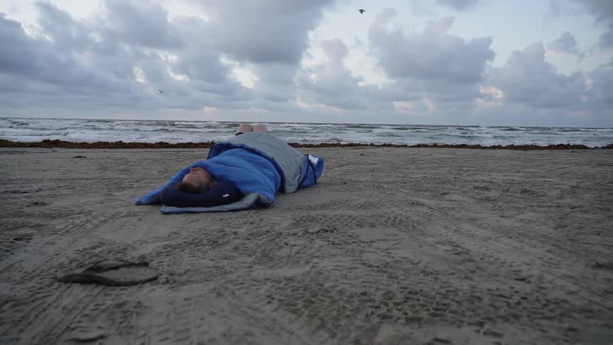 Man lays in a sleeping bag sleeping on the beach