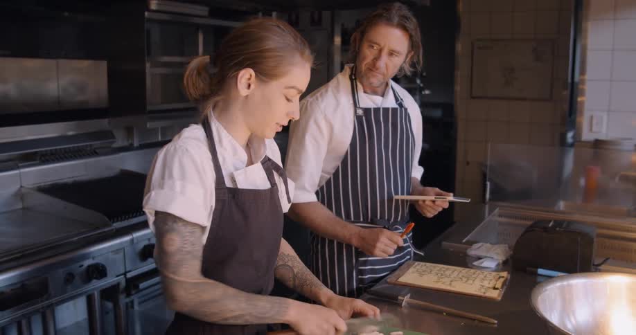 Man and woman talking while working in a kitchen and the woman is cutting mushrooms and the man is writing on a pad