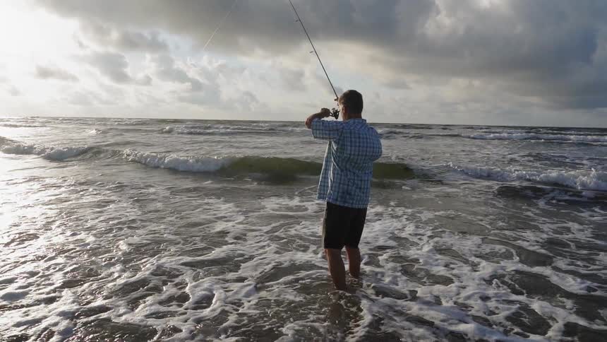 A man reels in his fishing line while standing in the surf