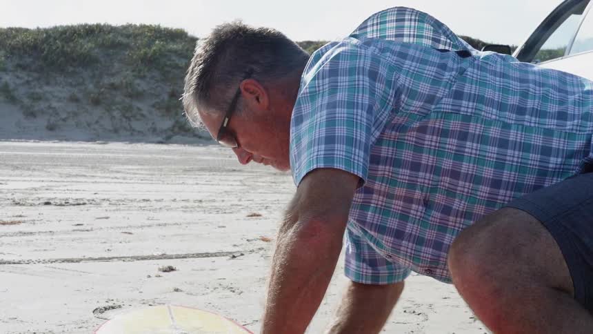 A man waxes a surfboard on a beach