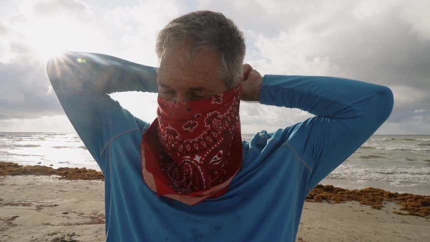Man puts a kerchief around his face as he stands on the beach