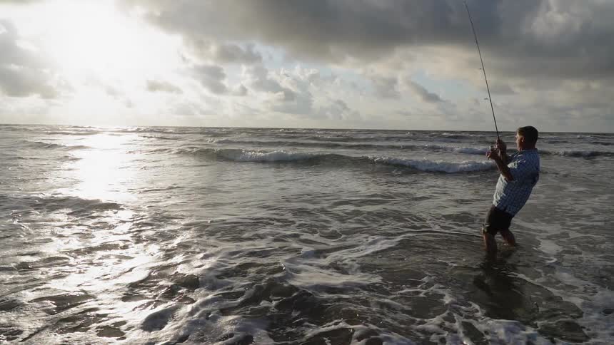 A man wades in ocean water while fishing on a beach