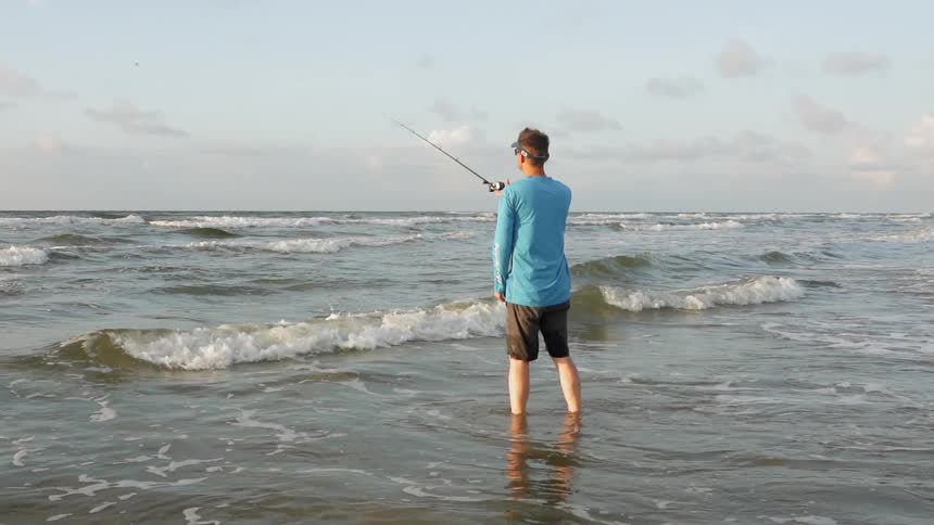A man wades in ocean water while fishing on a beach