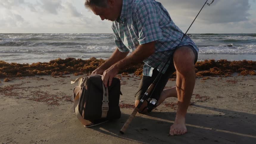 A man kneeling on the sand unzips a fishing gear bag